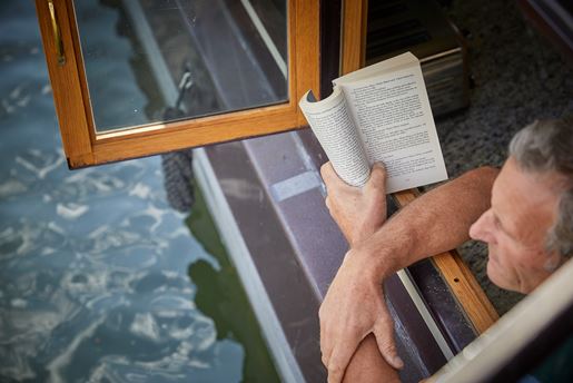 A man reading a book at the window of a canal boat.