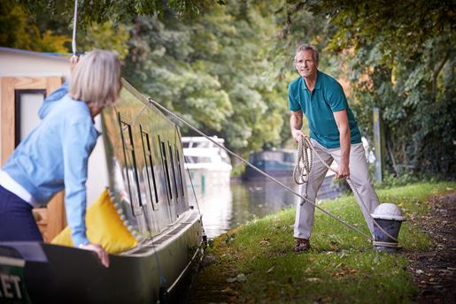 A man and woman look at each other as he moors a canal boat at the riverside and she watches from the boat, surrounded by greenery.