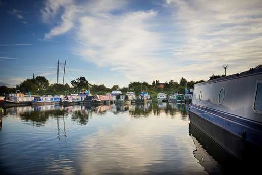 Moored canal boats on the riverside with reflective water and a bridge.