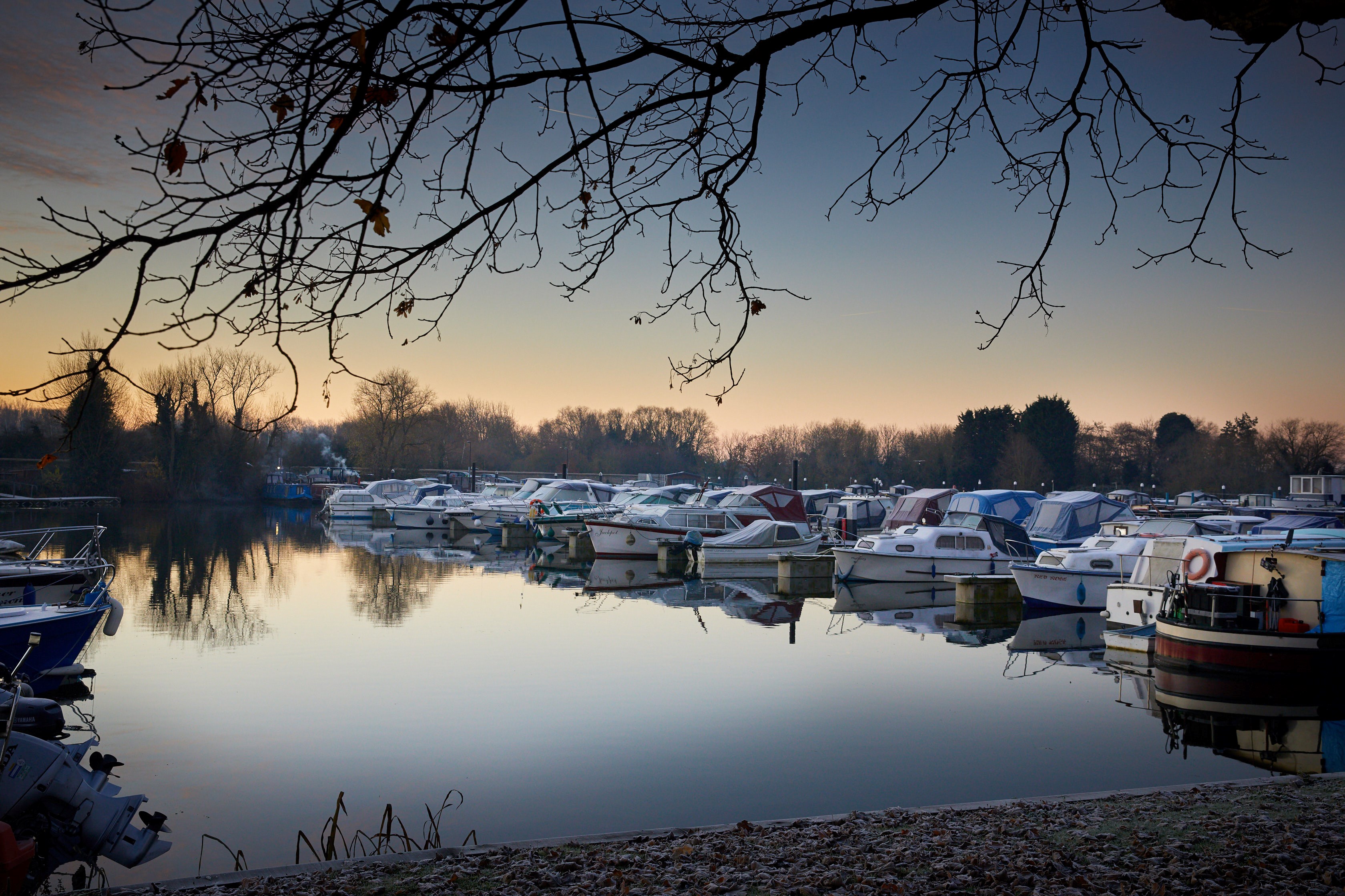Boats at the canal side at dusk in winter, framed by trees.